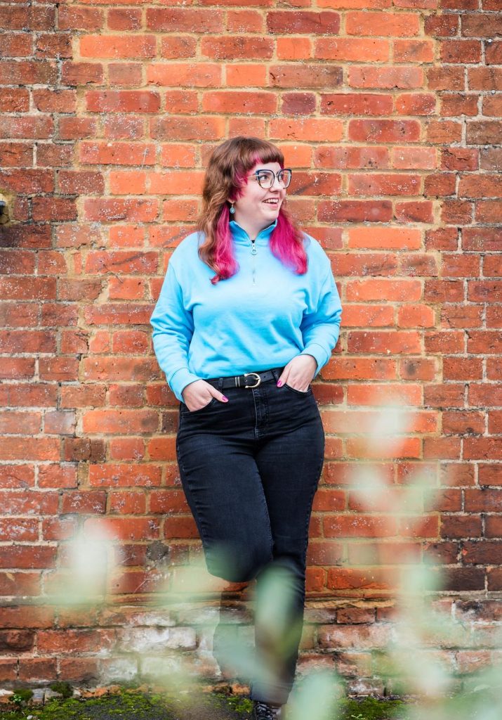 Photo of Emily Jones, founder of Two Shoes Creative. She has pink hair, big glasses, wears bright blue and is leaning up against a wall, smiling.